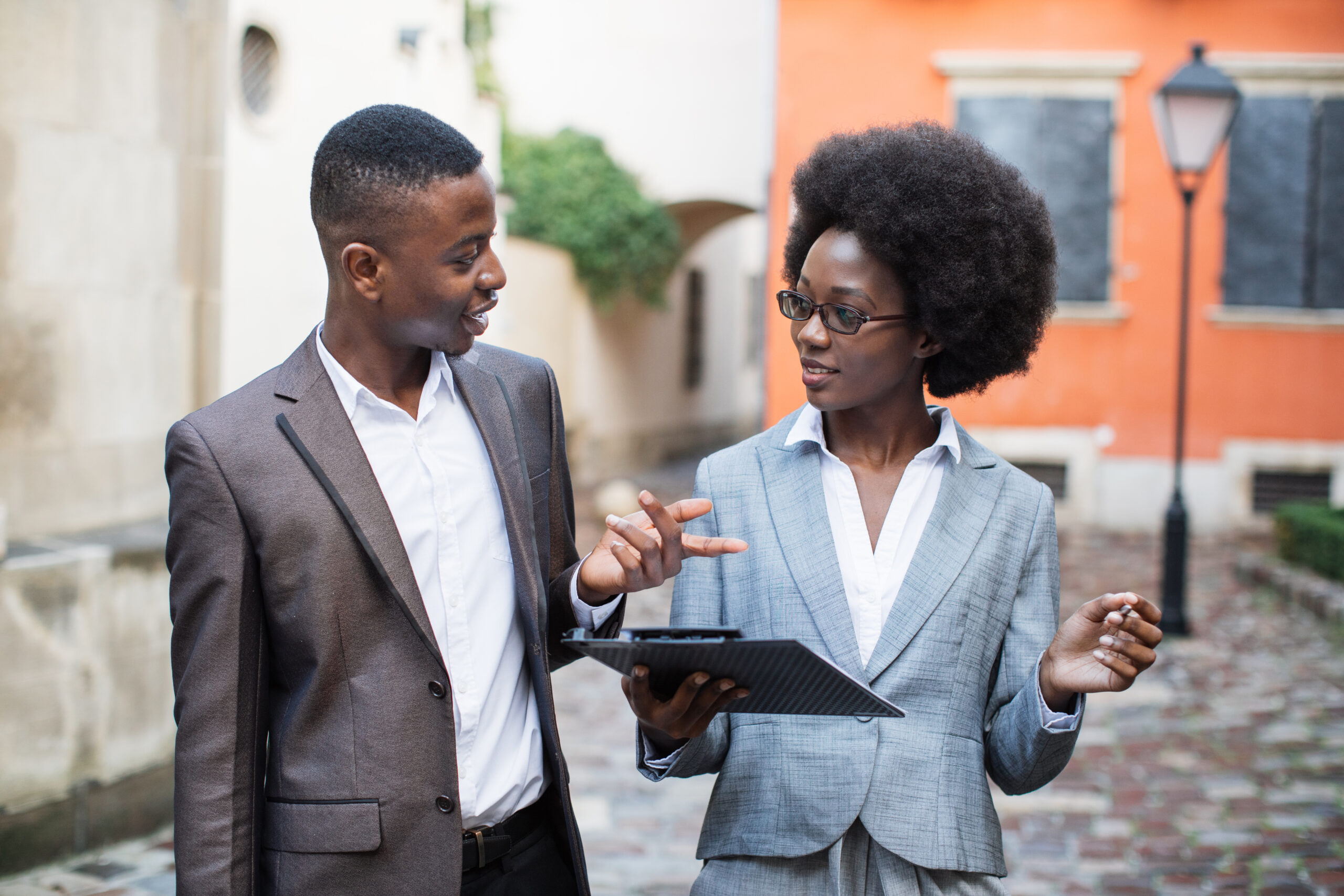 Charming woman with clipboard and handsome man with briefcase standing on street and communicating. Afro american business colleagues discussing some working issues outdoors.